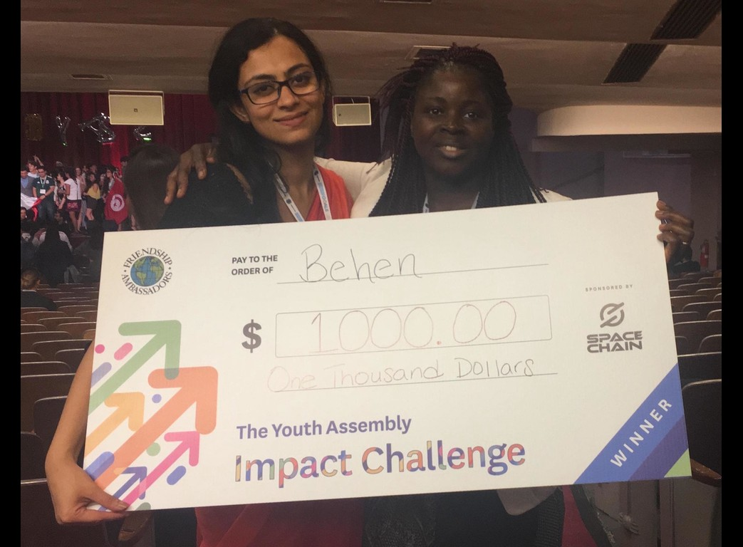 Two young women smile while holding a large ceremonial $1,000 check for winning The Youth Assembly Impact Challenge, in an auditorium with theater seating and a distant stage.